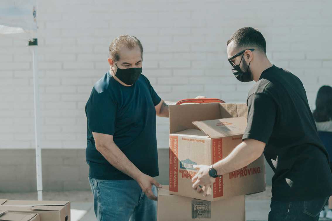 Two men wearing masks and stacking cardboard boxes